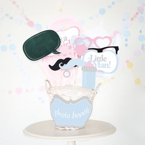May include: A white wicker basket filled with photo booth props for a baby shower. The props include a chalkboard speech bubble, a mustache, a pacifier, a baby bottle, glasses, and a sign that says "Little Man!" The basket is sitting on a white table.