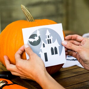 May include: A person is holding a paper stencil of a haunted house and a bat in front of an orange pumpkin. The stencil is printed on white paper with a gray background.