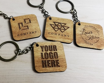 May include: Four wooden keychains with different designs. The keychains are square shaped and have a metal ring and chain. The designs include a geometric pattern, a logo with the text "COMPANY", and a logo with the text "Laser Kitchen" and the text "Let's Craft Your Idea" and "Since 2018". The fourth keychain has the text "YOUR LOGO HERE".