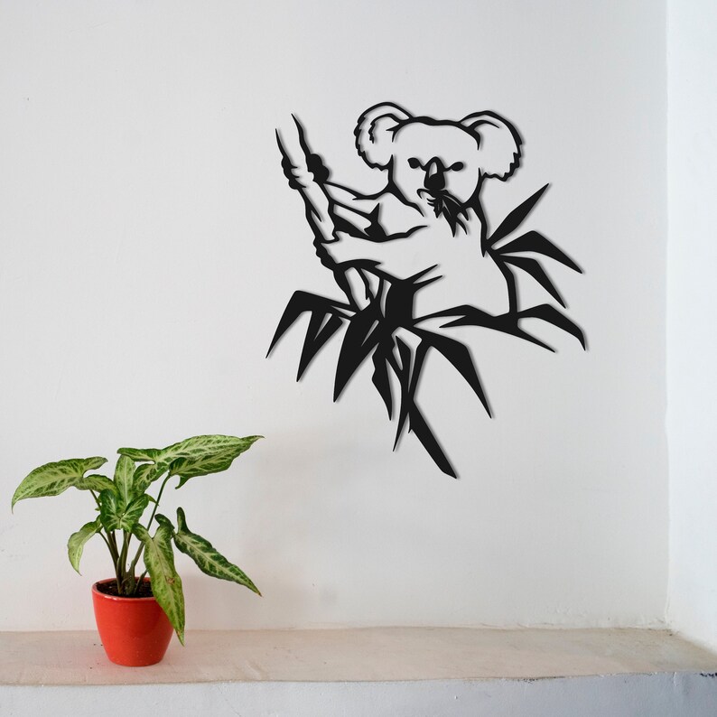May include: Black metal wall art featuring a koala bear sitting in a tree. The koala is in a relaxed pose, with its arms wrapped around a branch. The tree is stylized with leaves and branches.
