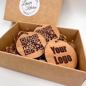 Può includere: Tre etichette rotonde in legno in una scatola marrone. Due etichette hanno un design a codice QR in bianco e nero. La terza etichetta ha il testo "Your Logo" inciso.