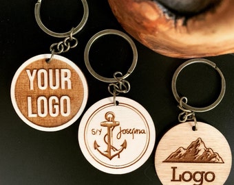 May include: Three wooden keychains with different designs. The first keychain has the text "YOUR LOGO" engraved on it. The second keychain has an anchor and the text "S/Y Josegma" engraved on it. The third keychain has a mountain range and the text "Logo" engraved on it.