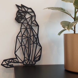 May include: Black geometric cat wall decor made of wood. The cat is in a sitting position with its tail curled around its legs. The cat is against a white wall with a potted plant in the background.