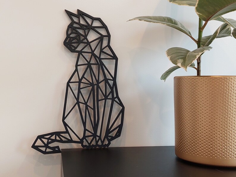 May include: Black geometric cat wall decor made of wood. The cat is in a sitting position with its tail curled around its legs. The cat is against a white wall with a potted plant in the background.