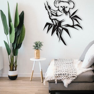 May include: Black metal wall art featuring a koala bear sitting in a bamboo tree. The art is displayed on a white wall in a living room setting with a couch and a side table.