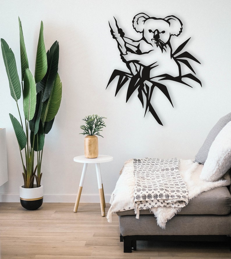 May include: Black metal wall art featuring a koala bear sitting in a bamboo tree. The art is displayed on a white wall in a living room setting with a couch and a side table.
