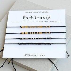 May include: Four black string bracelets with Morse code beads in gold, black, and silver are displayed on a white card. The card reads "FUCK TRUMP" in large letters and "MORSE CODE JEWELRY" above. The phrase ".GET LUCKY STYLE." is printed below.