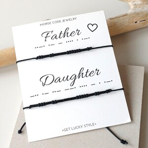 May include: Two black Morse code bracelets on a white card with the words "Father" and "Daughter". A heart symbol is above "Father". The card is on a neutral background with a wooden stick.