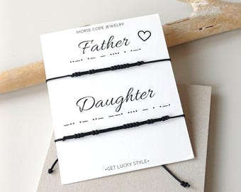 May include: Two black Morse code bracelets on a white card with the words "Father" and "Daughter". A heart symbol is above "Father". The card is on a neutral background with a wooden stick.
