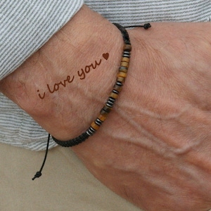Peut inclure: Un bracelet en cordon noir avec des perles brunes et argentées. Le bracelet a un petit charme en forme de cœur et les mots "i love you" écrits sur le poignet.