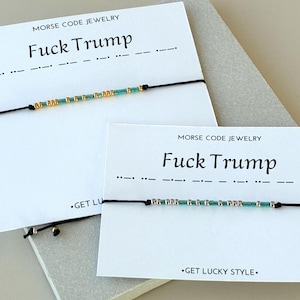 May include: Two bracelets with black cords and small beads in gold and teal colors. The bracelets are displayed on white cards with the text "MORSE CODE JEWELRY" and "Fuck Trump". The phrase is also represented in Morse code.