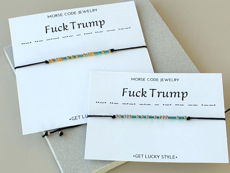 May include: Two bracelets with black cords and small beads in gold and teal colors. The bracelets are displayed on white cards with the text "MORSE CODE JEWELRY" and "Fuck Trump". The phrase is also represented in Morse code.