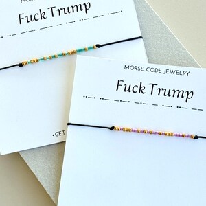 May include: Two bracelets with black cords and beaded Morse code spelling "Fuck Trump." The beads are gold, teal, and pink. The bracelets are displayed on cards with the text "MORSE CODE JEWELRY" and "Fuck Trump."