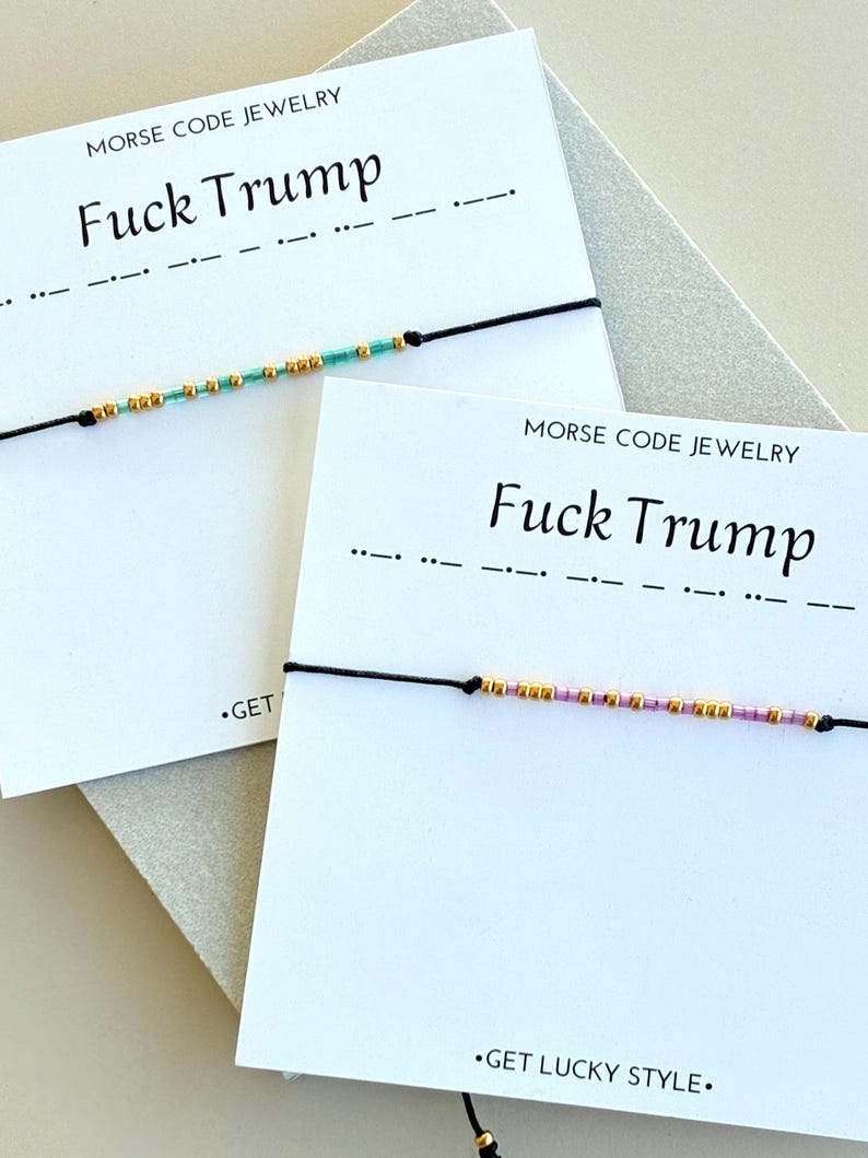 May include: Two bracelets with black cords and beaded Morse code spelling "Fuck Trump." The beads are gold, teal, and pink. The bracelets are displayed on cards with the text "MORSE CODE JEWELRY" and "Fuck Trump."