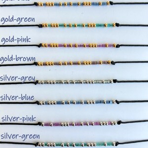 May include: A collection of beaded bracelets in various color combinations, including gold and silver beads with grey, blue, green, pink, and brown accents. Each bracelet is strung on a black cord.