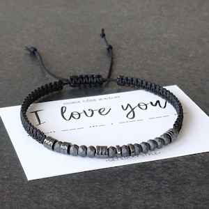 May include: Black braided bracelet with gray beads and metal accents. The bracelet is on a white card that says "I love you". The bracelet has an adjustable sliding knot closure.