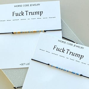 May include: Two black string bracelets with Morse code beads. The cards behind the bracelets read "MORSE CODE JEWELRY" and "Fuck Trump". One bracelet has gold and silver beads, the other has blue and silver beads.