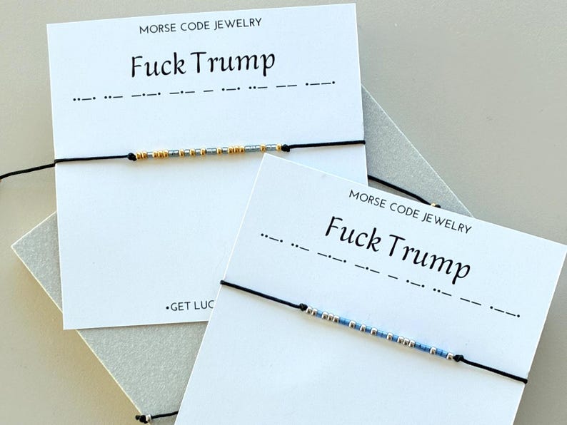 May include: Two black string bracelets with Morse code beads. The cards behind the bracelets read "MORSE CODE JEWELRY" and "Fuck Trump". One bracelet has gold and silver beads, the other has blue and silver beads.