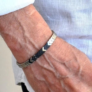 May include: A silver and black bracelet with a chevron pattern. The bracelet is made of metal and features interlocking arrow-shaped segments. The bracelet is worn on a wrist, with a white shirt visible in the background.
