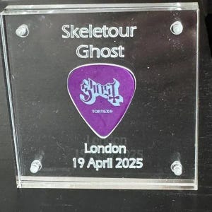 May include: A square clear acrylic display case containing a purple guitar pick with the Ghost logo and Tortex text. The case features white printed text reading Skeletour Ghost, London, and 19 April 2025, secured by four silver metal corner screws.