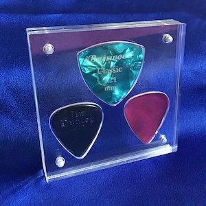 May include: A clear acrylic display case containing three guitar picks. The picks are triangular in shape and made from different materials. The top pick is turquoise blue with the text "Barewood Classic 1.21 mm". The left pick is black with the text "Jim Dunlop". The right pick is a deep magenta colour.