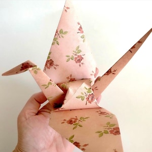 May include: A paper origami crane in a light pink color with a floral pattern of red and green flowers. The crane is folded into a traditional bird shape, with wings, a head, and a tail.