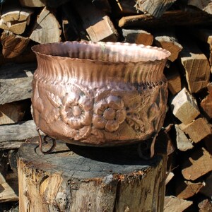 Könnte beinhalten: Ein Kupfertopf mit Blumenmuster steht auf einem Baumstumpf. Der Topf hat eine abgerundete Form und drei Beine. Der Hintergrund ist ein Stapel Brennholz.
