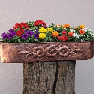 Könnte beinhalten: Ein kupferfarbener rechteckiger Pflanzkübel gefüllt mit bunten Blumen. Der Pflanzkübel hat ein Blumendesign auf der Vorderseite und steht auf einem verwitterten Holzstumpf. Die Blumen umfassen rote, violette, gelbe, orange und weiße Blüten.