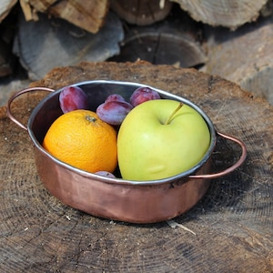 May include: A copper bowl with a handle on each side, filled with fruit. The bowl contains a green apple, an orange, and several purple plums.