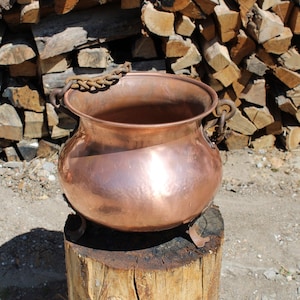 May include: A large copper pot with a chain handle and three legs sits on a wooden stump. The pot is shiny and has a hammered texture. The background is a pile of stacked firewood.