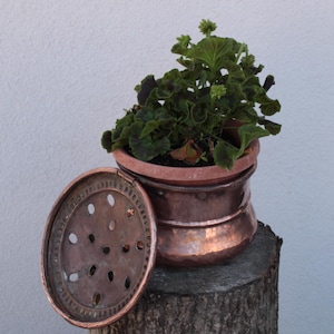 Op de afbeelding: Een gehamerde koperen pot met een bijpassend rond deksel met een geperforeerd ontwerp. De pot bevat een kleine groene plant in een terracotta-kleurige binnenbak. De koperen pot heeft een metallic afwerking en een ronde, brede vorm.