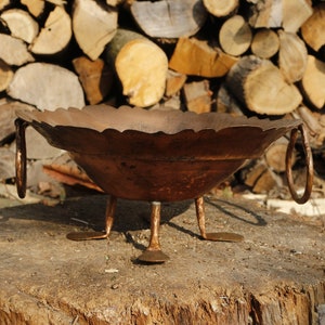 Puede incluir: Un cuenco de cobre con acabado martillado y tres patas se asienta sobre un tocón de árbol. El cuenco tiene un borde festoneado y dos asas.