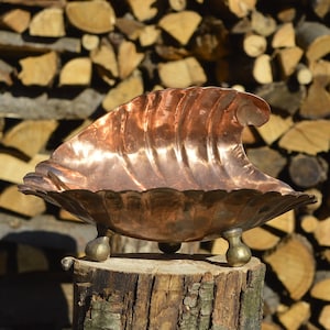 Op de afbeelding: Een koperen kom met een golvend ontwerp en drie poten staat op een boomstronk. De kom is glanzend en reflecteert het licht. De achtergrond is een stapel brandhout.