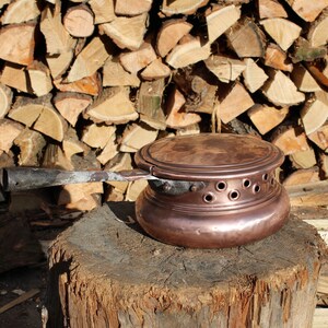 Puede incluir: Una olla de cobre con tapa y mango de madera se encuentra sobre un tronco de árbol. La olla tiene agujeros alrededor de la base y está rodeada de una pila de leña.