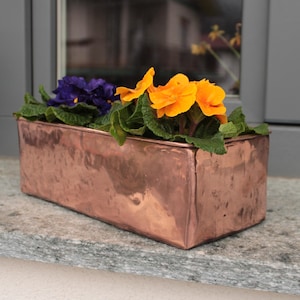 May include: A rectangular copper planter filled with vibrant flowers. The planter holds a mix of purple and bright orange blooms with green foliage. The planter sits on a stone surface near a window.