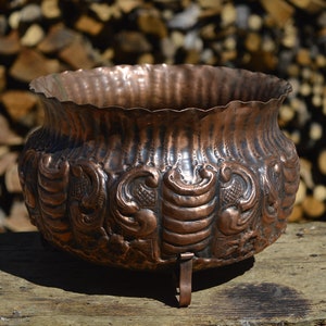 May include: A large, copper planter with an ornate, embossed design. The planter has three legs and is sitting on a wooden surface.