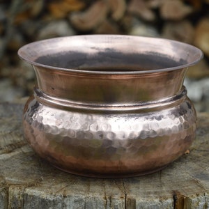 Pode incluir: Um vaso de cobre martelado com fundo arredondado e borda alargada. O vaso tem um aspecto rústico e envelhecido.
