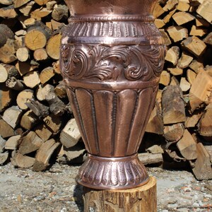 May include: An antique copper vase with detailed floral patterns and a hammered finish. The vase has a flared top and a decorative base. It is set on a wooden log, with a background of stacked logs, showcasing its craftsmanship.