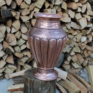 May include: A copper vase with an ornate design sits on a wooden stump. The vase has a wide base and a narrow neck, and it is decorated with a pattern of raised lines and swirls. The vase is a beautiful example of traditional craftsmanship.