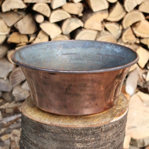 May include: A copper bowl with a slightly flared rim sits on a tree stump. The bowl has a patina of age and is slightly tarnished. The background is a pile of stacked firewood.