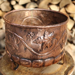 Può includere: Un vaso di rame con un designo a vite e foglie. Il vaso ha tre gambe ed è appoggiato su una superficie di legno.