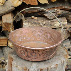 May include: A copper bowl with a floral design and a metal handle. The bowl is sitting on a wooden stump.