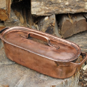 May include: A vintage copper roasting pan with a lid and two handles. The pan is oval shaped and has a hammered finish. It is sitting on a wooden stump.