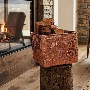 May include: A square, hammered copper basket with two side handles, filled with several pieces of split firewood. The basket is resting on top of a cylindrical, rough-textured wooden log stump, positioned in front of a stone fireplace.
