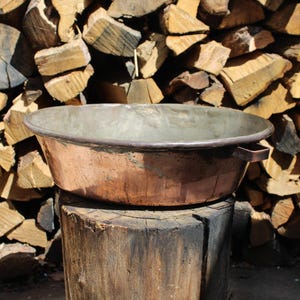 Op de afbeelding: Een verweerde koperen waskom met een breed, ondiep ontwerp, rustend op een houten stronk. De waskom heeft een donkere, verouderde patina en kleine handvatten. Een achtergrond van opgestapeld brandhout.