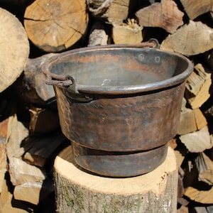 May include: A rustic, antique copper cauldron with a hammered texture, featuring a rounded body and a sturdy handle. The cauldron is set on a wooden stump against a backdrop of stacked firewood, showcasing a vintage aesthetic.