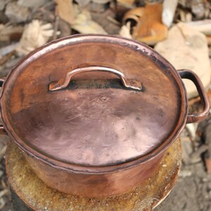 May include: A copper pot with a lid and two handles. The pot is sitting on a wooden surface.