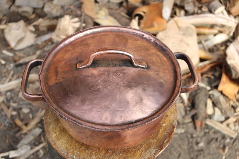 May include: A copper pot with a lid and two handles. The pot is sitting on a wooden surface.