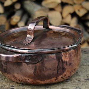 May include: A vintage copper pot with a lid and a handle. The pot is sitting on a wooden surface.