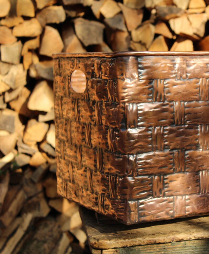 Puede incluir: Una cesta de almacenamiento de cobre tejido de forma cuadrada con un asa en el lateral. La cesta est&aacute; sobre una superficie de madera.